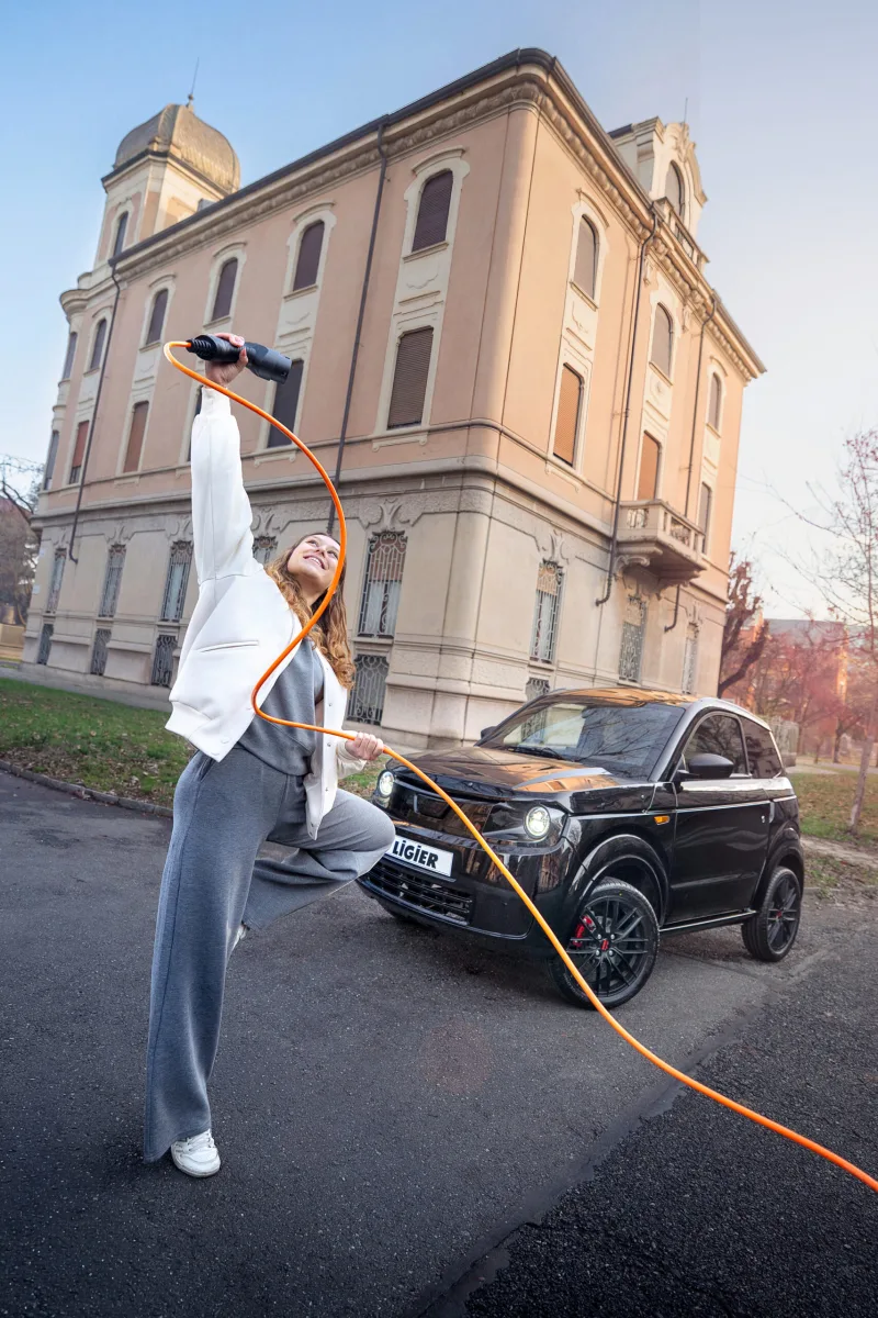 Ligier Myli noire garée en ville, câble de recharge orange tenu par une jeune femme